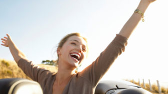 Young Woman Sits in the Back of a Convertible, Her Arms in the Air, Laughing With Joy