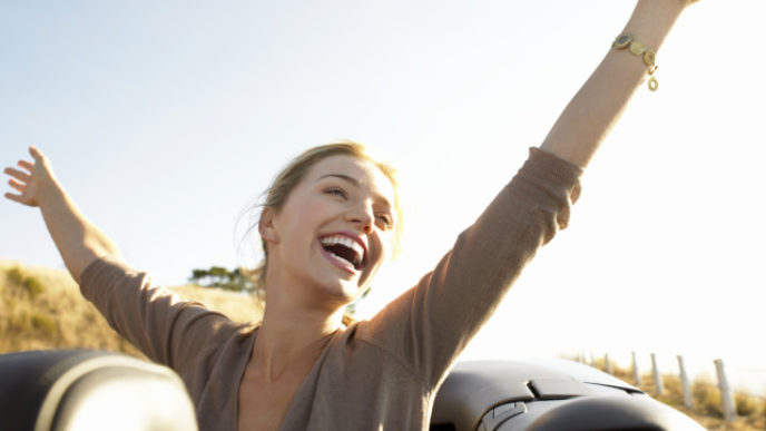 Young Woman Sits in the Back of a Convertible, Her Arms in the Air, Laughing With Joy