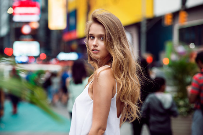 Beautiful blonde sexy tourist girl walking in busy city street with long hair flying on the wind. Woman looking at camera outdoors wearing fashionable white t-shirt. New York City lifestyle photo.
