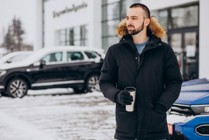 Handsome man in warm jacket standing by car covered with snow and drinking coffee