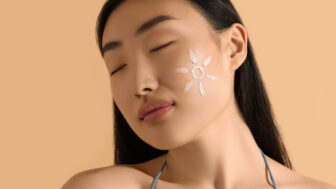 Beautiful young woman with sun protection cream on her face against beige background