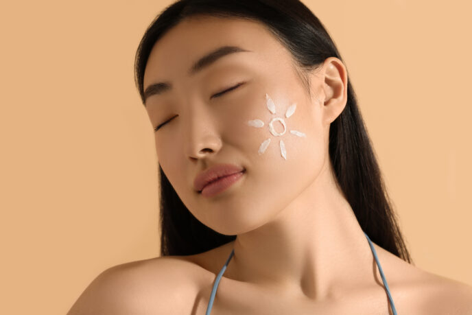 Beautiful young woman with sun protection cream on her face against beige background