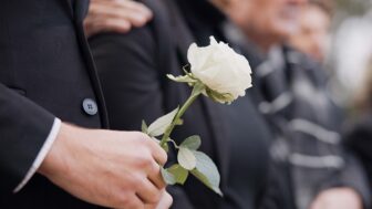 Hands, rose and a person at a funeral in a cemetery in grief while mourning loss at a memorial service. Death, flower and an adult in a suit at a graveyard in a crowd for an outdoor burial closeup