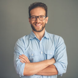 Handsome young businessman in shirt and eyeglasses is looking at camera and smiling while standing with crossed arms on gray background