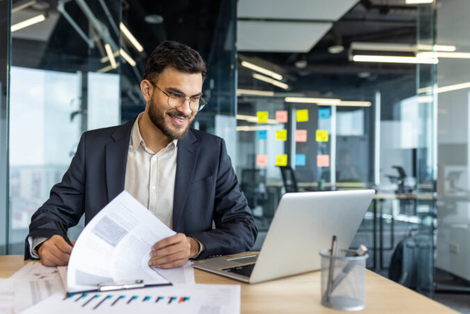 A businessman smiles as he works through documents on his desk next to a laptop computer.