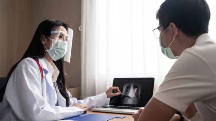 Female Doctor explain x ray chest result in computer laptop to illness man patient in examination room in hospital. Female medic wear face shield and mask during covid pandemic to prevent infection.