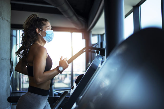 Sportswoman training on treadmill in gym and wearing face mask to protect herself against coronavirus during global pandemic of covid 19 virus.