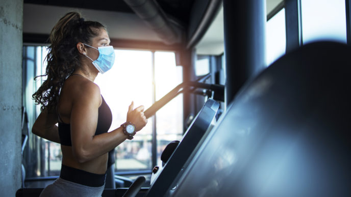 Sportswoman training on treadmill in gym and wearing face mask to protect herself against coronavirus during global pandemic of covid 19 virus.