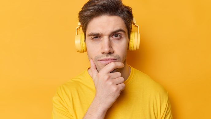 Horizontal shot of thoughtful man holds chin and looks seriously at camera listens favorite song via stereo headphones dressed in casual clothes poses against vivid yellow background. Let me think