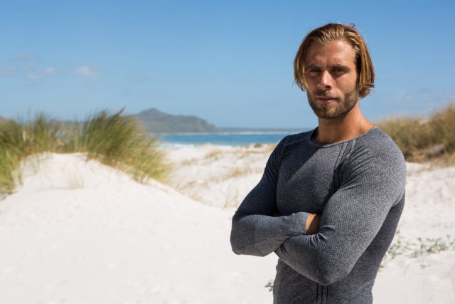 Portrait of confident athlete standing at beach against sky