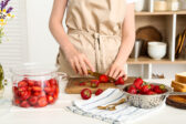 Woman cutting fresh berries for preparing sweet strawberry jam at table in kitchen