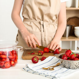 Woman cutting fresh berries for preparing sweet strawberry jam at table in kitchen