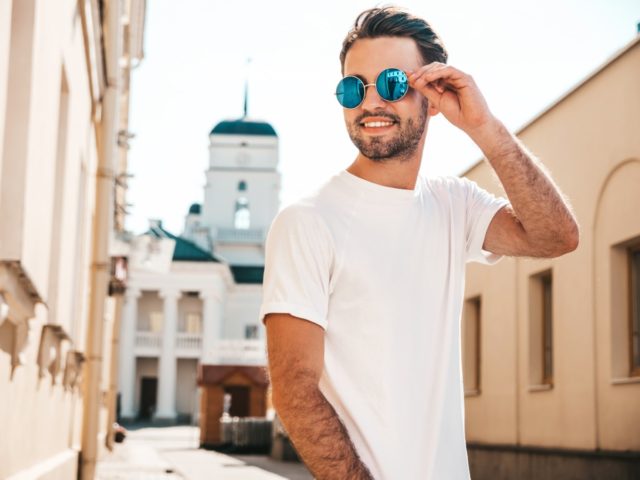 Portrait of handsome smiling stylish hipster lambersexual model.Man dressed in white T shirt. Fashion male posing on the street background in round sunglasses