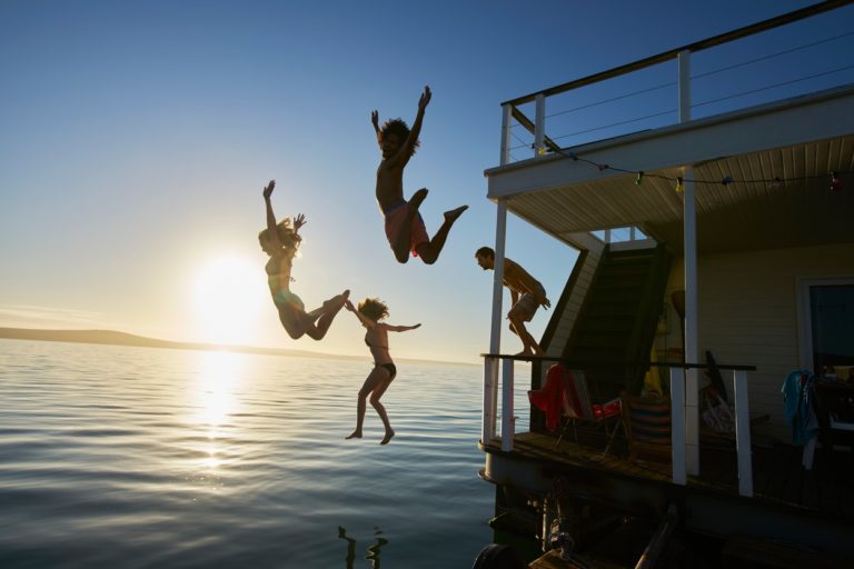 Young adult friends jumping off summer houseboat into sunset ocean