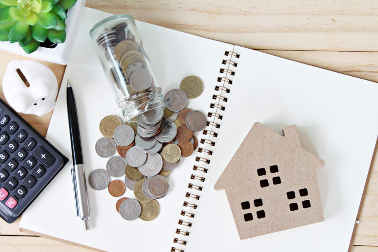 Flat lay or top view of wood house model and coins scattered from glass jar on open blank notebook paper