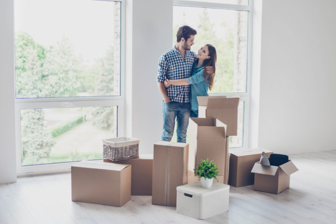 Successful young couple is moving to new nice place and embracing, around are carton boxes with their belongings. The room is very light and bright, they are wearing casual outfit