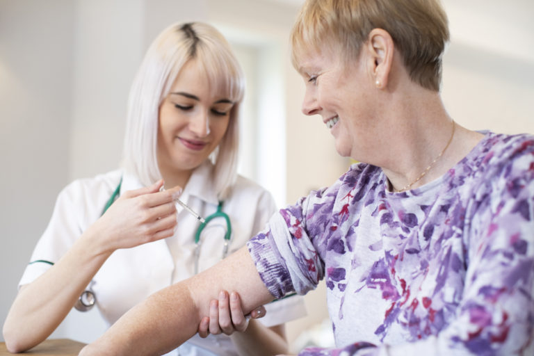 Female Nurse Giving Senior Woman Injection In Arm With Syringe