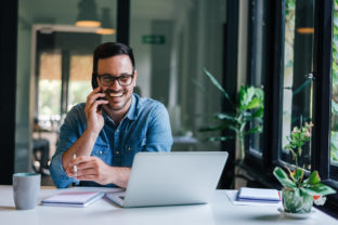 Portrait of young smiling cheerful entrepreneur in casual office making phone call while working with laptop