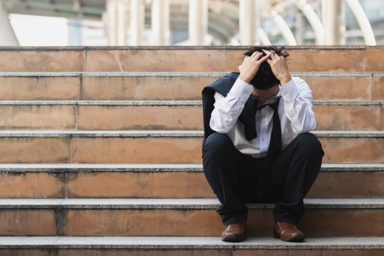 Upset stressed young Asian business man in suit with hands on head sitting on stairs. Unemployment and layoff concept.