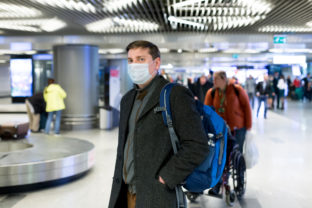 Young European man in gray coat, protective disposable medical mask in airport. Afraid of dangerous N CoV 2019 influenza coronavirus mutated and spreading in China. Blue backpack, suitcase on wheels