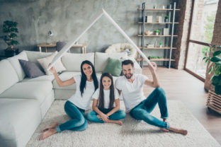 Portrait of nice attractive cheerful family wearing casual white t shirts jeans sitting on carpet floor holding in hand roof investment at industrial loft style interior living room cozy comfort