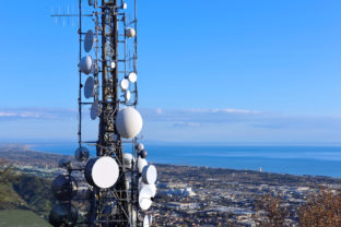 Aerial view of a telecommunication tower, satellite dish, network repeaters, base transceiver. Antenna for wireless network. Cellular station for smart city connect and coastline as background.