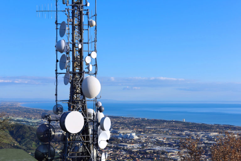 Aerial view of a telecommunication tower, satellite dish, network repeaters, base transceiver. Antenna for wireless network. Cellular station for smart city connect and coastline as background.