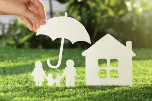 Woman holding paper umbrella over cutout of family and house on fresh grass, closeup. Life insurance concept