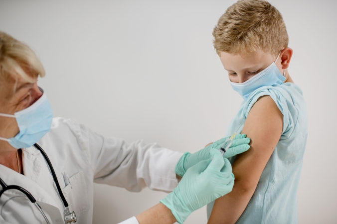 Pediatrician makes vaccination to cute Caucasian boy. They are wearing a protective face masks.