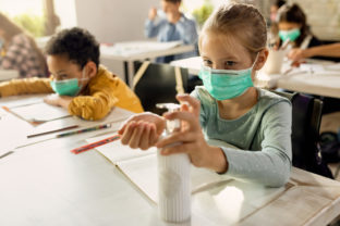 Elementary student wearing protective face mask and disinfecting her hands in the classroom.