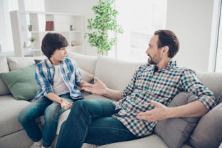 Portrait of two nice attractive friendly guys dad and pre teen son sitting on couch discussing psychology generation problems in light white modern style interior living room