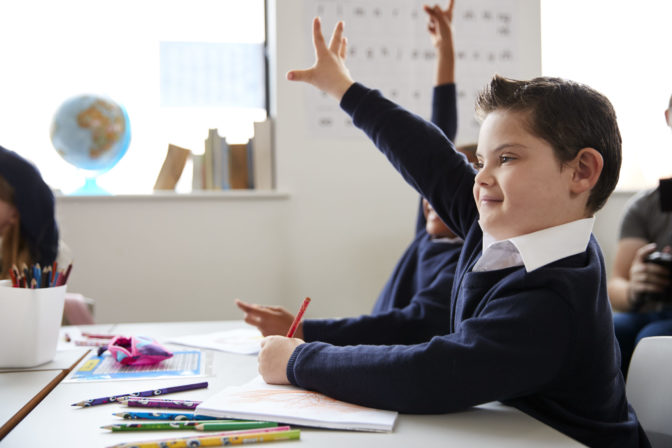 Schoolboy with Down syndrome sitting at a desk raising his hand in a primary school class, close up, side view