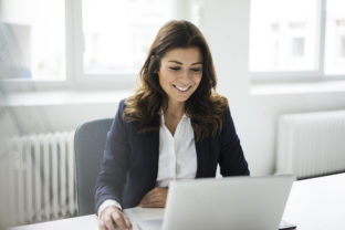 Portrait of smiling businesswoman sitting at desk in the office working on laptop