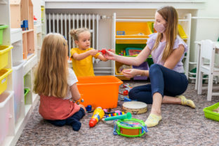 Young female therapist wearing protective face mask playing with two toddler girls during occupational child therapy.