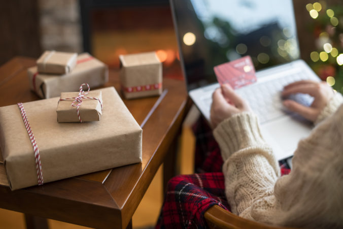 Christmas. Woman in sweater holding credit card using laptop for making order sitting at table with packaging gift near fireplace and christmas tree. Online shopping concept