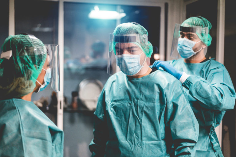 Doctors and nurse preparing to work in hospital for surgical operation during coronavirus pandemic outbreak - Medical workers getting dressed inside clinic - Focus on right man face