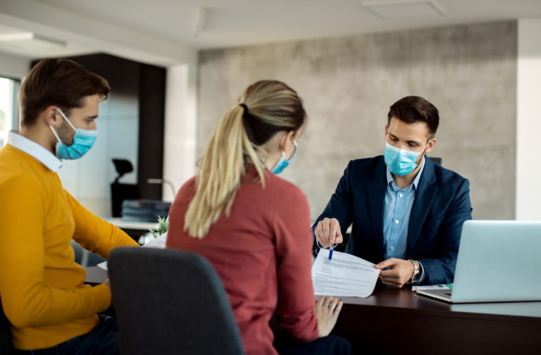 Insurance agent with face mask explaining terms of a contract to a couple during the meeting.