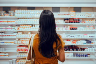 Rear view of young woman with bag standing against shelf in pharmacy searching for medicine