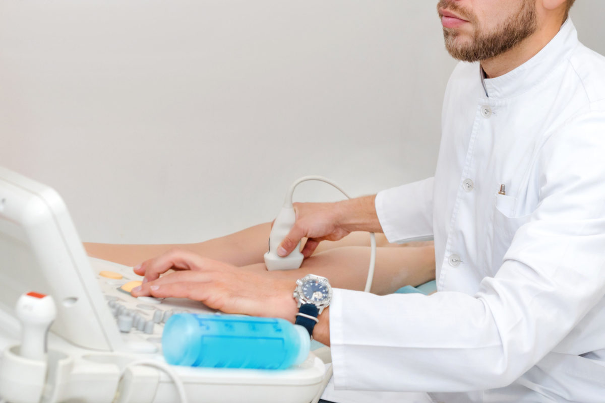 Doctor in white uniform with diagnostic equipment performs an ultrasound using a device on the legs of a female patient. Surgeon man, phlebologist working in moder clinic. Medical tools in hospital.
