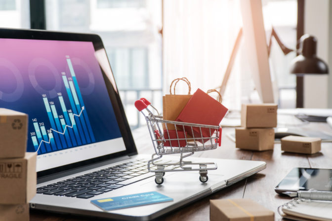 Shopping bags in shopping cart and credit card on laptop with paper boxes on table and sales data economic growth graph on screen, buying and selling services online on network, online shopping and e commerce concept