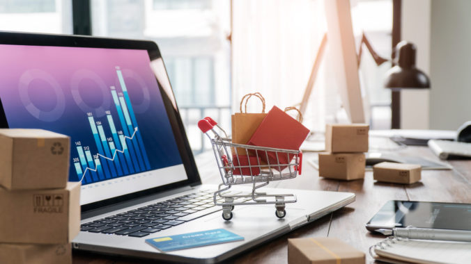 Shopping bags in shopping cart and credit card on laptop with paper boxes on table and sales data economic growth graph on screen, buying and selling services online on network, online shopping and e commerce concept