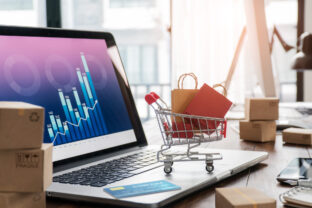 Shopping bags in shopping cart and credit card on laptop with paper boxes on table and sales data economic growth graph on screen, buying and selling services online on network, online shopping and e commerce concept