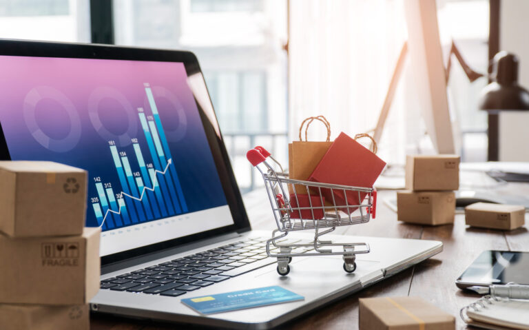 Shopping bags in shopping cart and credit card on laptop with paper boxes on table and sales data economic growth graph on screen, buying and selling services online on network, online shopping and e commerce concept