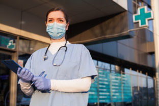 Young female doctor standing in front of healthcare facility, wearing protective face mask and PPE equipment, holding medical patient clipboard, COVID 19 pandemic crisis