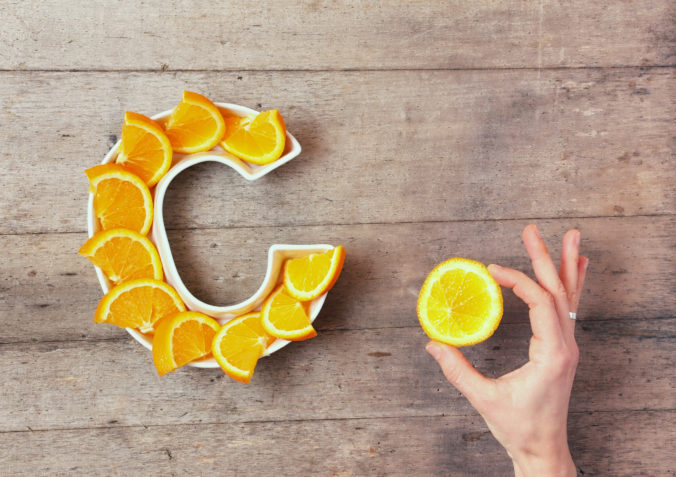 Vitamin C or Ascorbic acid nutrient in food concept. Plate in shape of letter C with orange slices and woman&#039;s hand with citrus making sign OK on wooden background. Flat lay or top view.