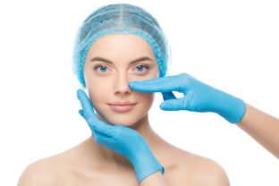 Young woman ready for rhinoplasty, doctor in blue gloves touching her nose, isolated on white background