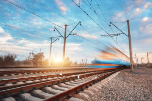 High speed blue passenger train in motion on railroad at sunset. Blurred commuter train. Railway station against colorful sky. Railroad travel, railway tourism. Rural industrial landscape. Vintage