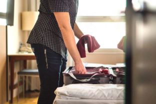 Man packing suitcase for vacation. Person putting clothes to baggagge in hotel room or home bedroom. Traveler with open luggage on bed.