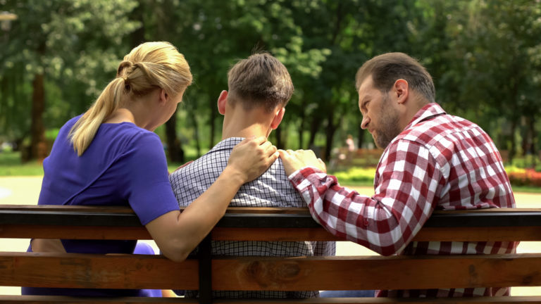 Caring mother and dad supporting sad teen son sitting on bench i