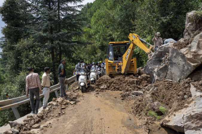 Scooter riders rush past an earthmover clearing a road of a big rock that came down with mud and plant debris in Dharmsala, Himachal Pradesh state, India, Sunday, Aug. 21, 2022. Landslides and flooding over the last three days killed at least three dozen people in this Himalayan state, an official government release said Sunday